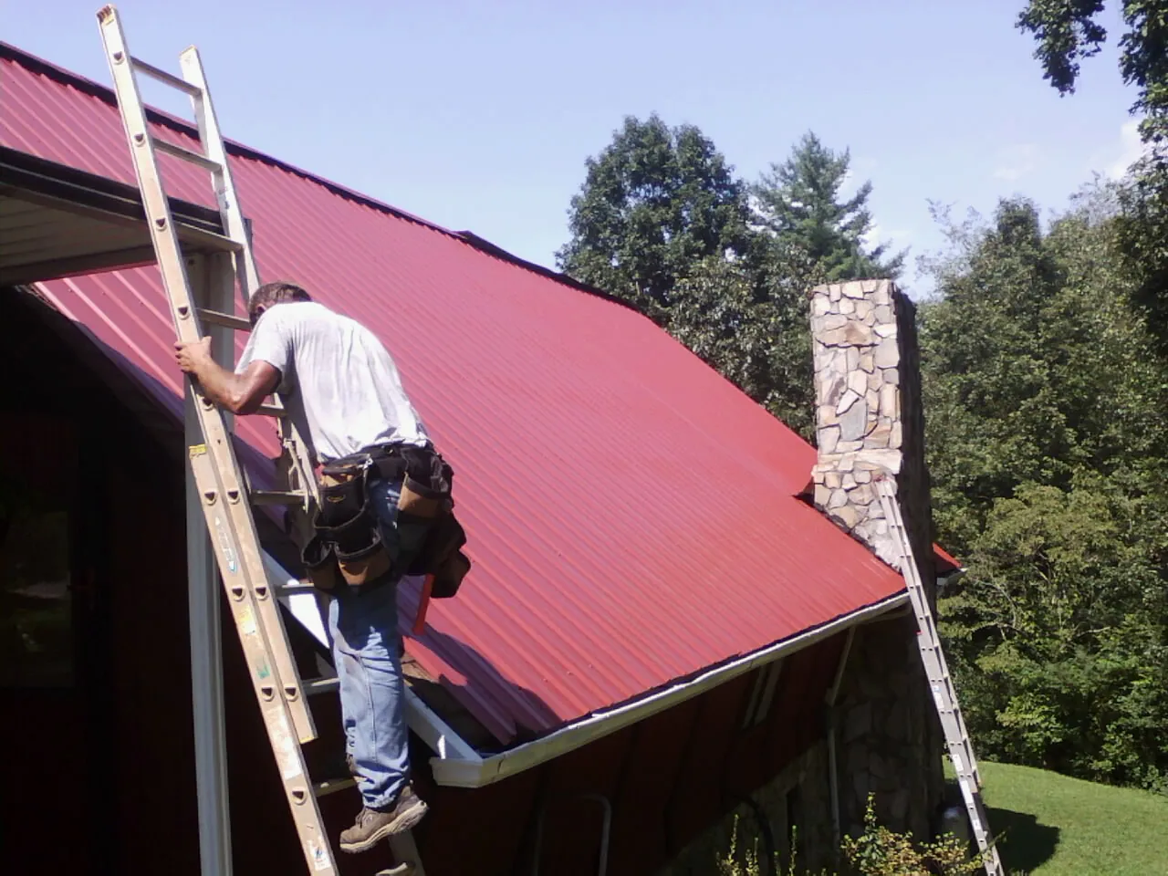 Roofer on a ladder beside a red metal roof and stone chimney — Soffit & Fascia Repair by Roofing Brothers in Hohenwald